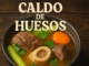 Caldo de huesos con carne, zanahoria y apio en tazón oscuro sobre mesa rústica. Ideal para recetas tradicionales y contenido de bienestar.