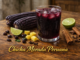 Vaso de chicha morada peruana con hielo, maíz morado, canela, piña y limón sobre mesa rústica con texto dorado.